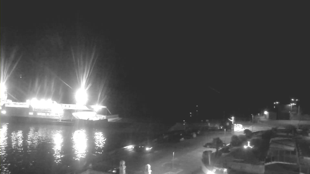 A dark waterfront scene at night, featuring a brightly lit boat docked on the left. The water reflects the lights from the boat and other sources. To the right, there is a partially visible parking area with a few vehicles and faint outlines of structures. The sky is mostly dark with minimal illumination from the surroundings.