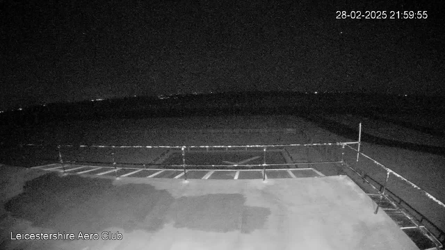 Black and white nocturnal image of an empty rooftop at Leicestershire Aero Club. The scene is mostly dark, with some faint distant lights visible in the background. There is a railing in the foreground and a flat surface below it, possibly a landing area or tarmac. The time stamp in the corner reads 28-02-2025 at 21:59:55.