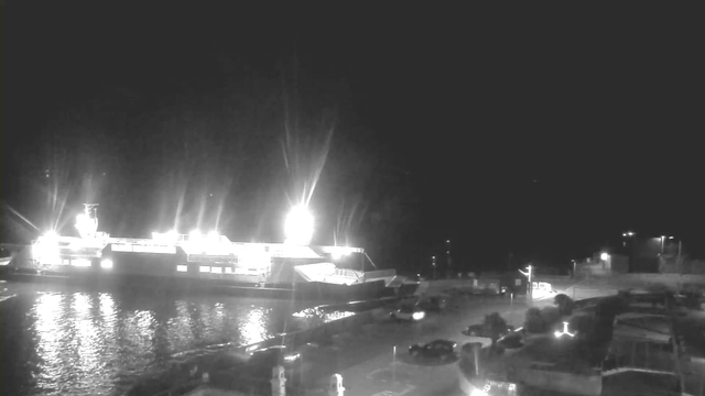 A brightly lit ferry is docked at night, casting reflections on the calm water. The ferry has multiple lights illuminating its structure, particularly at the front. In the foreground, there is a parking area with a few parked vehicles and a visible road leading away from the dock. The scene is mostly dark, highlighting the lights of the ferry and nearby buildings.