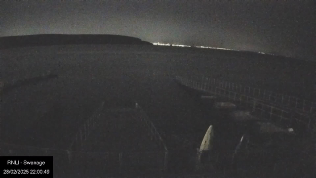 A dark image of a shoreline at night, featuring a calm sea extending towards a distant landmass. In the foreground, there are structures that may be docks or piers, partially illuminated by faint lights. The background shows a faint outline of hills and a sparse line of lights along the horizon, suggesting a small town or city. The overall mood is serene and quiet, capturing a night scene by the water.