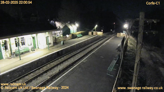 A dimly lit railway station platform at night. The scene includes a stone building with multiple windows and a green bench in front. There is a sign that reads "WAY OUT" positioned on the platform. The area to the right shows additional green benches and a wooden fence marking the exit. The railway tracks are visible, running through the center of the image, flanked by gravel and concrete paths. Soft lighting from a nearby lamp illuminates the area, creating a quiet and solitary atmosphere.