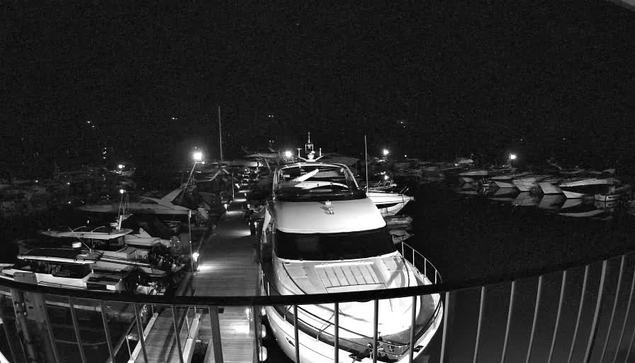 A nighttime scene at a marina, featuring several boats docked along a wooden walkway. The area is illuminated by various lights reflecting off the water, creating a peaceful atmosphere. Some boats are large, with white hulls visible in the foreground, while others are smaller and scattered throughout the harbor. Dark water surrounds the dock and boats, while the background is filled with more vessels, partially obscured in shadow.