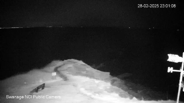 A dark seaside view captured by a webcam, showing gentle waves lapping against a rocky shoreline. There is a visible pathway leading toward the water, with a small sign partially illuminated. To the right, a weather vane stands out against the night sky, with the ocean stretching into the darkness. Some distant lights are faintly visible on the horizon, suggesting the presence of land or boats. The image timestamp indicates it is 23:01:08 on February 28, 2025.