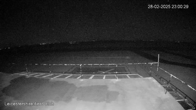 A dimly lit scene showing an airport tarmac at night. The foreground features a railing, with several white lines marking parking spots or runway sections. In the distance, faint lights can be seen, suggesting buildings or vehicles beyond the tarmac. The sky is mostly dark, indicating it is nighttime. A timestamp in the upper right corner shows "28-02-2025 23:00:29." The bottom left corner has the text "Leicestershire Aero Club."