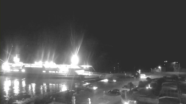 A nighttime scene showing a large ferry docked at a port, illuminated by bright lights. The water reflects the lights, creating a shimmering effect. In the foreground, several parked cars can be seen, with streetlights illuminating the area. The overall atmosphere is quiet and serene, typical of a late-night setting.