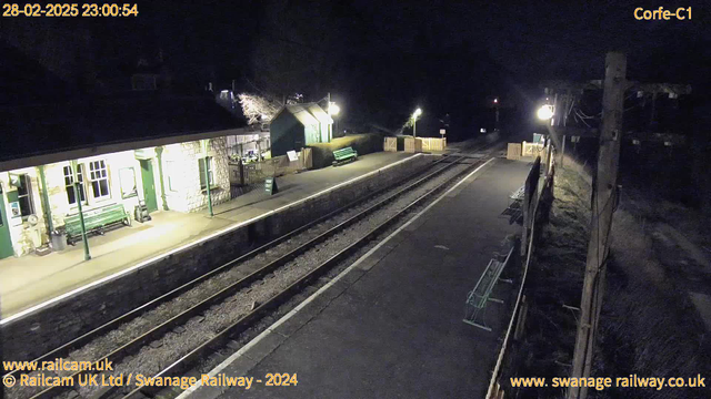 A dimly lit railway platform at night. The platform features a traditional stone building with large windows and a green door. There are several green benches along the platform and a wooden fence on one side. A sign reading "WAY OUT" is visible, indicating an exit. Two sets of railway tracks run beneath the platform, leading away into darkness. A few faint lights illuminate the area, casting soft shadows. The time displayed shows 23:00:54 on February 28, 2025.