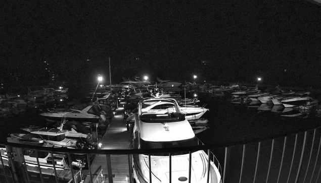 A nighttime scene at a marina, featuring numerous boats docked in the water. The image is predominantly in black and white, with some scattered lights illuminating the boats and the surrounding area. The foreground shows a railing and the top of a boat, while the marina extends into the distance, filled with various vessels. The atmosphere appears calm and serene, with reflections of lights on the water's surface.