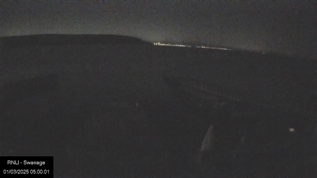 A dark scene captured from a webcam, showing a faint outline of hills or coastal land in the background with a dim horizon. The top half of the image features a night sky with a very low light level, while the lower part shows a silhouette of what appears to be a boat or dock area at the foreground. There are no prominent details visible, and the scene appears grainy due to low visibility. A timestamp in the bottom left corner indicates the date and time of the image.