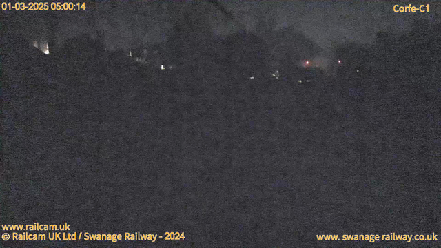 A dark, low-light scene captured by a webcam. The image displays blurred outlines of trees and structures in the background, with faint lights visible, possibly from distant buildings or vehicles. The overall atmosphere is dim and lacks distinct features, creating a mostly shadowy view. A timestamp in the top left corner indicates the date and time as March 1, 2025, at 05:00:14. Branding from the Railcam UK and Swanage Railway appears in the corners.