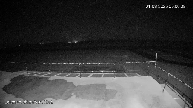 A dimly lit scene from a webcam at Leicestershire Aero Club shows the edge of a runway during nighttime. The foreground features a flat surface with some obscured markings, while a low fence runs along the side. In the background, multiple faint lights are visible, suggesting distant structures or vehicles. The date and time in the corner indicate it is early morning on March 1, 2025.