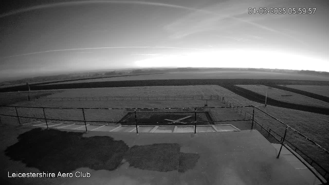 A black and white image from a webcam showing an open field with a runway at the Leicestershire Aero Club. The sky appears to be lightening as the sun rises, with faint streaks and clouds visible. The foreground features a large flat area, possibly a tarmac or concrete surface, with a marking resembling an "X" indicating a landing spot. A railing runs along the edge of the surface, with grass areas visible in the background. The date and time displayed are March 1, 2025, at 05:59:57.