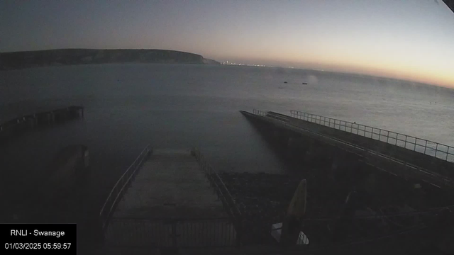 A dimly lit coastal scene at dawn, featuring a calm sea under a pale blue sky transitioning to orange and pink near the horizon. In the foreground, a wooden jetty extends into the water, while another jetty is visible on the right. There are small boats scattered on the water's surface. The landscape includes a distant shoreline with cliffs, and the time stamp indicates early morning.