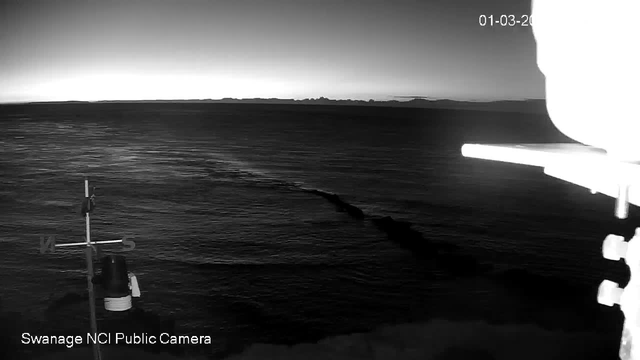 A black-and-white image of a calm sea under a clear sky. The horizon is visible in the background, with faint outlines of distant land. In the foreground, there is a navigational marker or pole with directional indicators. The time and date are displayed in the top right corner, with the text "Swanage NCI Public Camera" at the bottom. The water surface has light reflections, suggesting early morning or dusk.