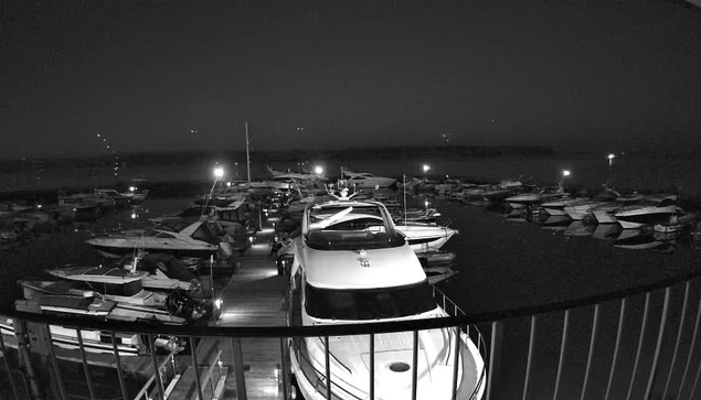 A nighttime scene of a marina filled with various boats docked along a wooden pier. The image is predominantly dark, with some dim lighting illuminating parts of the boats and the water. Several boats are visible, with some showing details like cabins and covers. The still water reflects faint light from the surrounding area. In the background, an indistinct horizon is visible, suggesting the presence of land.