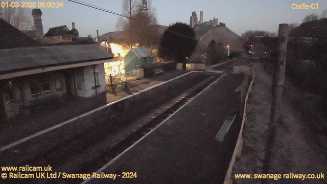 A dimly lit railway station scene at dawn. In the foreground, there are railway tracks with wooden sleepers and gravel on either side. To the left, a partially covered building architecture with a slanted roof is visible, indicating the station's structure. In the background, there is a green structure resembling a shed or small building. Beyond it, a hill rises with several tall, rugged stone structures that may be ruins or a castle. The atmosphere is quiet and serene, with soft light illuminating the area from the structure on the left.