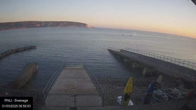 A coastal scene showing calm water with a distant view of cliffs in the background. In the foreground, there is a wooden ramp leading down to the water, bordered by a metal railing. To the right, there are two concrete piers extending into the water, one with a railing and the other unrailed. A yellow kayak and a blue boat are visible on the ground nearby. The sky is lightening with dawn colors, and the water is mostly smooth with a few small boats scattered in the distance.