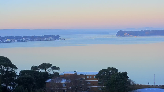 A wide view of a tranquil body of water at dawn, reflecting soft pastel colors of blue and pink. In the foreground, there are trees and a low building with a snow-covered roof. On the horizon, a small town with boats and distant hills can be seen along the shoreline, creating a serene landscape.