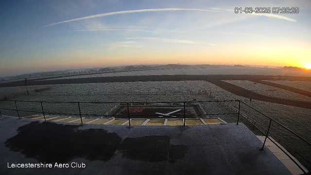 A panoramic view from a webcam at Leicestershire Aero Club, showing a frosty landscape at sunrise. In the foreground, there is a flat rooftop area with a railing. A small white runway marking is visible on the ground. The horizon features a soft gradient of colors from pale orange to blue as the sun rises. The background includes fields with a light frost, and a few distant trees are visible under a clear sky. The time stamp indicates it's early morning on March 1, 2025.