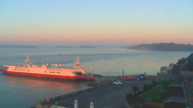 A ferry is docked in a calm harbor during early morning light. The sky is a gradient of soft colors, transitioning from light blue to peach. The water is smooth, and in the distance, there are small islands partially shrouded in mist. In the foreground, a white car is parked near the water's edge, and a few scattered structures can be seen along the shoreline. There are some palm trees and greenery near the waterfront, and a few distant boats are visible on the water.