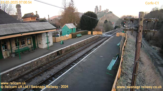 An image of Corfe Castle Railway Station taken from a webcam, showing empty train tracks lined with gravel. The station features a green and white building with a slanted roof, several green benches along the platform, and a wooden fence to the side. In the background, rolling hills contain the ruins of Corfe Castle. The scene is bathed in early morning light, and there is a mix of bare trees and shrubs. A wooden utility pole stands nearby with wires extending from it. The image is timestamped 07:00:54 on March 1, 2025.