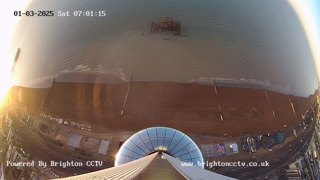 Aerial view from a webcam at sunrise, capturing a beach scene. The sandy shoreline curves along the bottom of the image, with several large wooden structures partially submerged in the water. Gentle waves lap at the shore, and the sea stretches into the horizon. A large circular structure, possibly part of an amusement park, is visible in the foreground, along with other buildings and attractions. The sky is illuminated with soft, warm colors as the sun rises. The timestamp indicates it is Saturday, March 1, 2025, at 07:01:15.