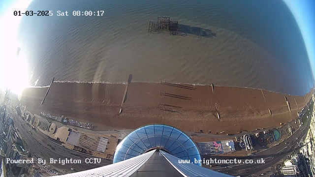 A panoramic view from a high vantage point overlooking a beach. The upper half of the image shows a clear blue sky. Below, the sandy beach stretches horizontally, with gentle waves lapping at the shore. Several wooden structures are visible in the water and along the beach. To the right, there are outlines of buildings and amusement facilities, while the left shows a line of parked vehicles. The scene is well-lit, indicating early morning hours. A circular glass dome is present at the bottom of the image, suggesting it is taken from an observation tower.