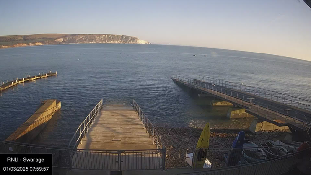 A peaceful seaside scene at sunrise, featuring calm waters and a rocky shore. There are two piers extending into the water, with a curved pier on the left and a straight pier on the right. Several small boats are moored in the distance, and a yellow kayak is visible on the beach. The background features a hilly coastline with trees. The sky is clear, transitioning from orange to blue, indicating early morning light.