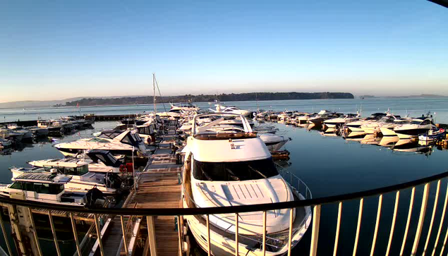 A scenic marina filled with numerous boats docked in calm waters. In the foreground, a large white yacht is visible, along with smaller boats surrounding it. The sky is clear and blue, reflecting soft morning light on the water's surface. In the background, there is a distant shoreline with trees, complementing the serene atmosphere of the harbor.