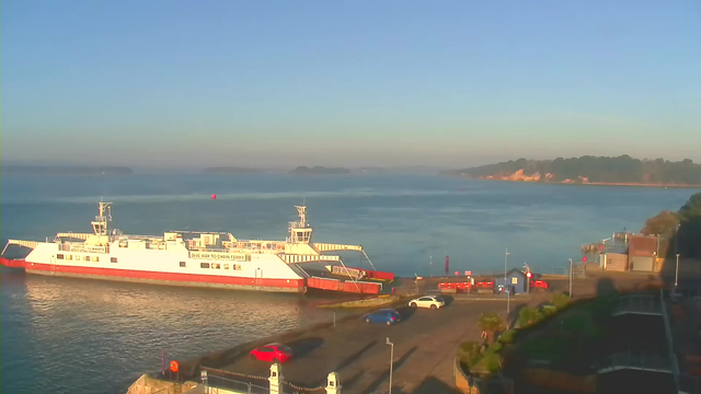 The image shows a calm waterfront scene during early morning. A large white ferry with orange accents is docked on the left side of the image. In the foreground, there are several parked cars, including a red one and a blue one. The setting is peaceful with clear skies, and the water reflects the soft morning light. In the background, there are trees and a shoreline with a faint outline of land visible in the distance.