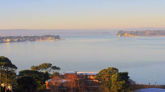 A tranquil view of a calm body of water at dawn, reflecting soft pastel colors in the sky. To the left, there is a small marina with several boats and a coastal settlement lined with trees. In the foreground, a building with a sloped roof and scattered trees partially obstruct the view of the water. The distant shoreline features a wooded area and a small island with a structure visible on it. Overall, the scene conveys a peaceful, serene atmosphere.