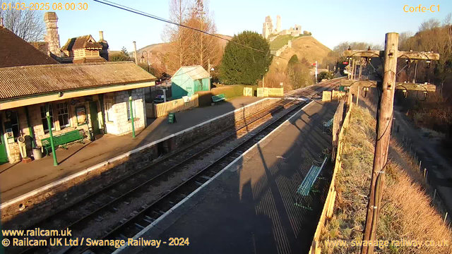 A railway station scene is depicted during the early morning. The platform, lined with benches, is bathed in soft sunlight. To the left, there is a station building made of stone featuring large windows and a sloped roof. A green structure resembling a shed is visible in the background, alongside a tall, leafy tree. In the distance, the silhouette of a castle atop a hill can be seen against the sky. The tracks lead off toward the right, with a telephone pole positioned on the edge of the platform, supporting various wires. There is a sign on the platform that reads "WAY OUT."