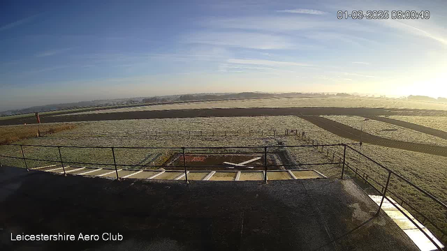 A view from a webcam shows a clear sky with some wispy clouds. The foreground features a runway with a red-and-white striped pole on the left. A large, grassy area is visible, with a prominent section marked by a white cross on a concrete pad, indicating a landing or takeoff area. In the distance, rolling hills are partially obscured by a light mist. The overall scene conveys a calm, early morning atmosphere.