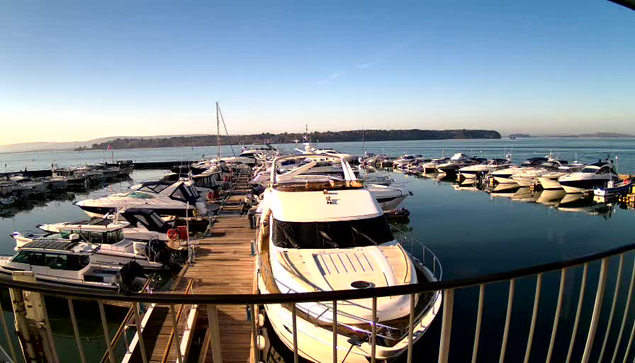 A marina scene featuring numerous boats docked in calm water. The foreground shows various yachts and smaller vessels moored along a wooden pier. In the background, the water reflects the blue sky and surrounding hills, with a few boats visible further out. The atmosphere appears peaceful and sunny, with clear visibility.