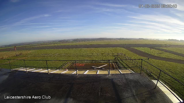 A clear view of a grassy airport runway under a blue sky with some clouds. In the foreground, there's a brown and white helipad marked with an "X." Surrounding the helipad are low fences. To the left, a tall red and white airport navigation tower is visible. The runway is mainly dark grey with patches of green grass alongside. The scene is well-lit, indicating daytime.