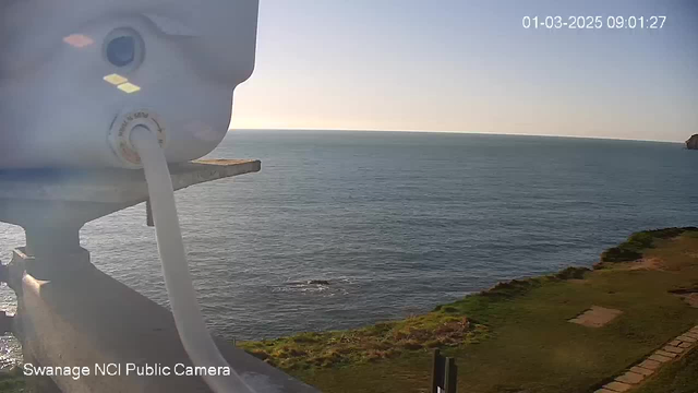 The image shows a view from a webcam overlooking the ocean. In the foreground, there is a white device mounted on a railing, with a cable extending from it. The background features a calm sea with gentle waves reflecting light from the sun, which is positioned low in the sky. The shoreline is grassy with some patches visible, and a rocky outcrop is seen on the right side of the image. The scene is bright and clear, indicating a sunny day. The date and time are displayed at the top right corner.