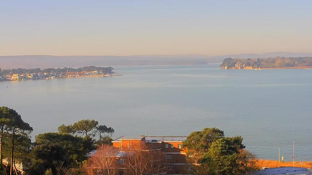 A tranquil waterfront view features calm waters with gentle ripples reflecting the soft light of dawn. In the distance, a marina is dotted with various boats and yachts, while the shoreline is lined with trees. On the foreground, a cluster of buildings with orange and brown hues is partially visible, surrounded by greenery. The landscape is framed by rolling hills in the background, creating a serene and picturesque scene.