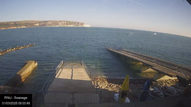 A sunny coastal scene featuring a calm sea with gentle waves. In the foreground, there's a concrete ramp leading down to the water, bordered by metal railings. To the right, a small yellow kayak is resting on the shore among pebbles. In the distance, white cliffs rise along the shoreline under a clear blue sky. A boat is visible on the horizon, and a pier extends into the water on the left.