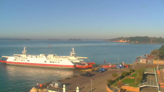 A ferry is docked at a harbor on a clear day with blue skies. The ferry is white with a red stripe and has the words "Chain Ferry" visible. In the foreground, there are parked cars and a walkway beside a grassy area with some palm trees. The water is calm, reflecting the sunlight, and a rocky shoreline is visible in the distance.