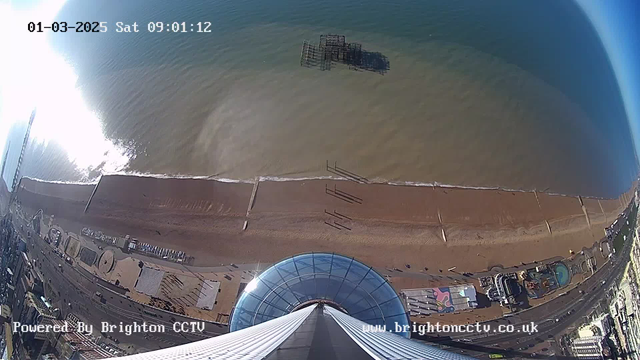 Aerial view of a beach and coastline taken from a high point. The image shows brown sandy beach with a gentle wave lapping at the shore. In the water, a partially submerged structure, likely remnants of a pier, is visible. The ocean appears calm with a blue-green hue and reflections of sunlight sparkling on the surface. In the foreground, a circular glass structure with a domed roof is positioned, below which there are some visible patterns on the ground. The bottom right corner shows portions of an amusement park with colorful rides and installations. The image indicates strong sunlight shining from the top left corner.