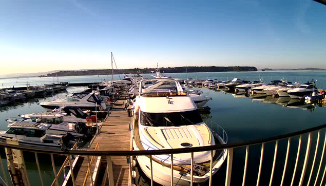 A view of a marina on a clear day. Numerous boats are docked along a wooden pier. In the foreground, a white yacht is prominently positioned, with several other boats visible in the water around it. The water is calm and reflects the blue sky above, while a distant shoreline can be seen in the background. The overall scene suggests a tranquil and sunny day by the water.