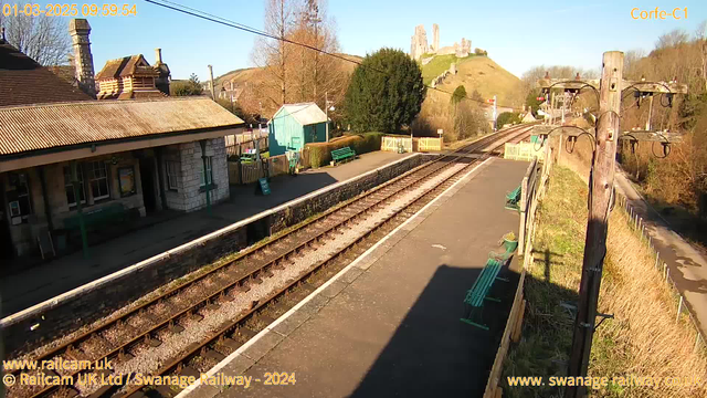 A view of a railway station in the foreground with two sets of railway tracks running parallel to each other. On the left, there is a stone building with a sloped roof, featuring a chimney and multiple windows. Nearby, there are several green benches along the platform. In the background, a vivid green hill rises, topped by the ruins of a castle. The sky is clear and blue, indicating a sunny day. A wooden utility pole with electrical lines stands on the right side of the image.