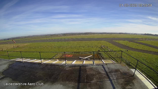 A wide view of an airport runway and grassy area under a bright blue sky with wispy clouds. In the foreground, a helipad with a white cross marking is visible, surrounded by a railing. To the left, a windsock indicates wind direction and speed, while several runways intersect the grassy landscape. The scene appears clear and bright, suggesting a sunny day.