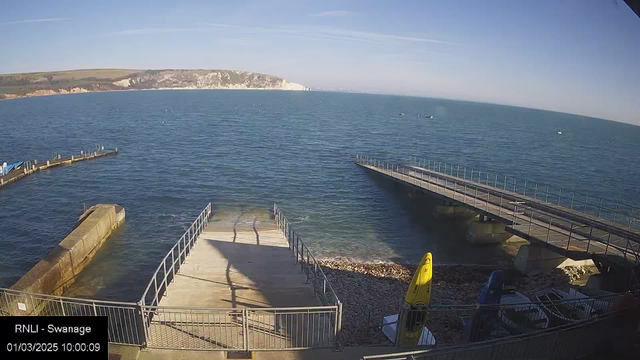 A coastal scene shows calm blue waters, with a rocky shoreline on the right and grassy hills in the background. There are two piers extending into the water: one on the left with a boat ramp and another wooden pier on the right. Several boats can be seen on the water. In the foreground, there are kayaks and a small boat resting on the ground. The sky is clear with a few wispy clouds. The timestamp on the bottom indicates the date is January 3, 2025, at 10:00 AM.