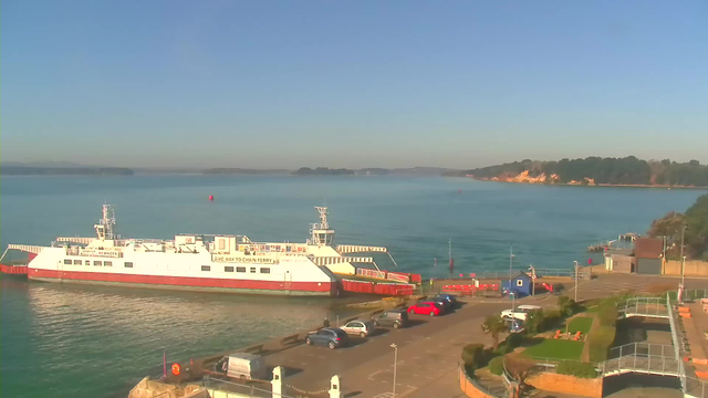 A ferry is docked at a calm waterfront under a clear blue sky. In the foreground, there are several parked cars and a pathway leading to the ferry terminal. The water is a vibrant blue with gentle ripples. In the background, green hills line the shore, and a few boats can be seen in the distance. The ferry has a white and red color scheme with signage indicating "Give Way to Chain Ferry."