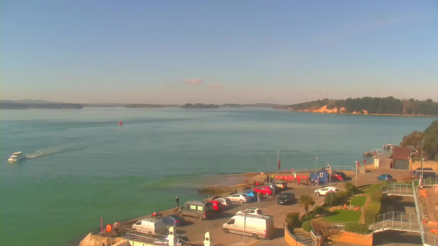 A tranquil waterfront scene with clear blue skies and calm waters. In the foreground, there is a parking area filled with vehicles, including red and white cars, vans, and a blue building. People are seen near the water's edge. A small boat is moving in the water, with a buoy visible nearby. In the background, green hills and sandy cliffs extend along the shoreline.