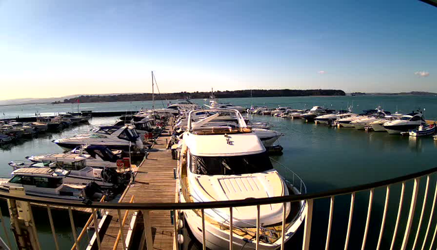 A scenic view of a marina filled with various boats docked at wooden piers. The foreground shows several white and blue yachts, along with smaller boats. The water is calm, reflecting the clear blue sky above, with a few clouds scattered. In the background, land can be seen beyond the marina, creating a peaceful coastal atmosphere.