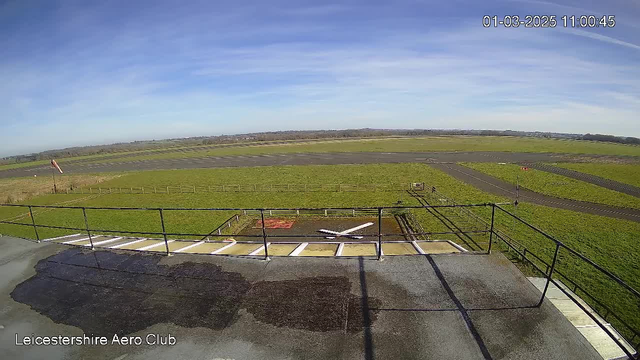 A wide view of an airport runway and surrounding landscape taken from an elevated position. In the foreground, there is a flat rooftop with a railing and some wet patches on the surface. Below, a small white aircraft symbol is visible on a grassy area. The background features a flat expanse of green grass, with a few rows of wooden fencing, leading to a paved runway marked with black tarmac. The sky is clear with few scattered clouds, indicating sunny weather.