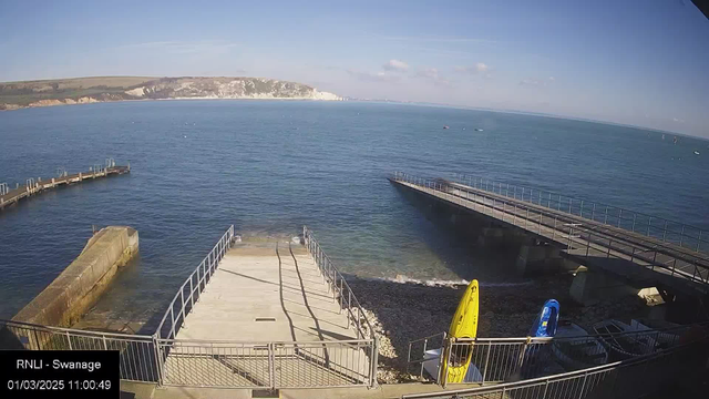 A clear view of a coastal scene, featuring calm blue water and sky. In the foreground, there is a wide concrete ramp leading down to the water, bordered by metal railings. On the right, two kayaks are tied up, one yellow and one blue, sitting beside a rocky beach. In the background, a wooden pier extends out into the water, with another pier visible further to the left. Light clouds are scattered in the sky, and cliffs can be seen in the distance, indicating a natural landscape. The timestamp in the bottom left corner shows the date and time.