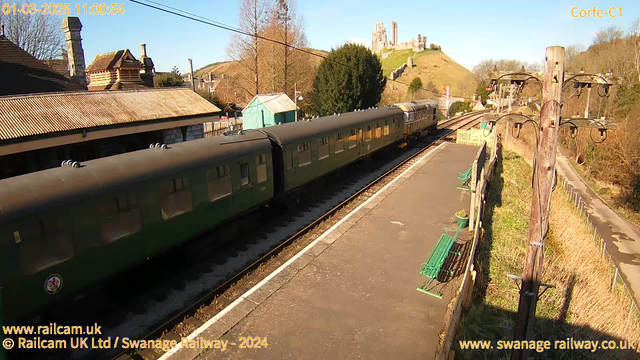 A railway station scene under a clear blue sky. A green train with multiple carriages is stopped on the tracks. In the background, there is a historic castle perched on a hill, surrounded by trees. The station features a traditional building with a sloped roof and intricate architectural details. There are wooden benches along the platform and a telegraph pole with wires running alongside the tracks. The overall atmosphere is bright and sunny, typical of a picturesque countryside setting.