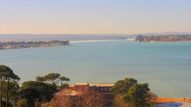 A scenic view of a calm, blue water body surrounded by a green shoreline. In the distance, small boats are docked along a waterfront lined with houses and trees. On the right, a small island with buildings and trees can be seen, while gentle hills rise in the background. The sky is clear with soft light illuminating the scene, indicating a pleasant day. In the foreground, there are some trees and a brick building partially visible.